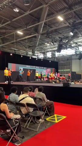 A stage performance is taking place inside a large indoor venue. The stage is set up with a black backdrop and a colorful screen displaying images. A group of performers, some of whom are young people, are on stage. Some are dressed in bright yellow and orange shirts. Others are in blue and pink shirts. Several performers are seated behind large, red drums, playing them with drumsticks. There's also a woman in a purple shirt standing on the left, also holding drumsticks and possibly leading the group. The audience is seated in chairs in front of the stage, with many people visible. The venue appears to be a convention center or exhibition hall, indicated by the industrial ceiling structure and lighting. The overall atmosphere suggests a cultural event or festival. There are signs visible on the backdrop, including one with the word "SAKURA".