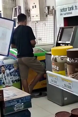 A person wearing a black t-shirt and tan shorts stands in front of a refrigerator in a store. The refrigerator has a picture of a soccer player on it. To the right of the refrigerator is an ATM machine with a screen. Jars of food products are stacked on a shelf in front of the ATM machine. The person is holding a phone in their hands and looking down at it. They appear to be interacting with the phone. The environment appears to be a retail store or convenience store.