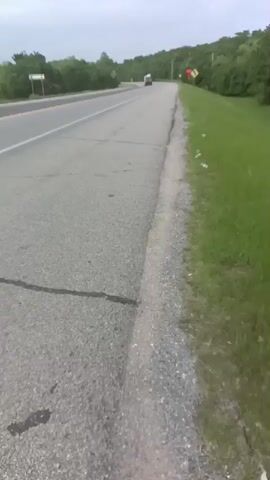 The video shows a two-lane rural road under an overcast sky. A truck pulls a trailer, followed by a box truck, both traveling away from the camera. Green foliage lines the sides of the road. A crack runs across the road's surface near the bottom of the frame. A small piece of trash is visible next to the road. A road sign is visible on the side of the road.