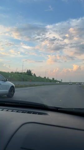 This video captures a view from inside a vehicle traveling on a highway. The focus is on a white Audi SUV in front, which is driving on the right lane. The SUV has tinted windows and black rims. The road is asphalt, with a concrete barrier on the left and a grassy shoulder with trees in the distance. Streetlights line the highway. The sky above is a mix of blue and dramatic, puffy clouds, with hints of pink and orange suggesting either sunrise or sunset. Other vehicles are visible in the distance on the highway. The dashboard of the vehicle from which the video is filmed is partially visible in the foreground. The overall impression is of a scenic drive under a dynamic sky.