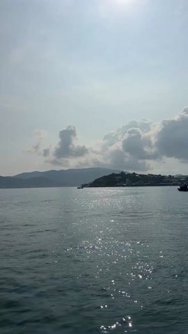 The video captures a serene seascape under a bright, partially cloudy sky. The sun glints off the water's surface, creating a sparkling effect. In the distance, a landmass with a small settlement is visible. Two boats are seen in the water, one further away and another closer to the viewer. The overall scene conveys a sense of tranquility and maritime activity.