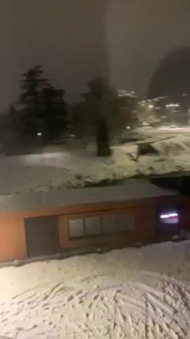 The video shows a snow-covered landscape at night, seen through a window. The ground is white with snow, and there are buildings, trees, and streetlights visible. A modern, low-rise building with orange siding is in the foreground, also covered with snow. Further in the distance are lit platforms and a circular area, presumably a roundabout, blanketed in snow. The scene is illuminated by streetlights.  There appears to be a "no" sign visible on the window due to reflection.