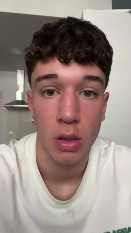 A close-up shot shows a young man in a white t-shirt looking directly at the camera. He has short, curly dark hair, fair skin, and green eyes. He sticks his tongue out, displaying it to the viewer. The background shows a clean kitchen area with a stainless steel range hood and white cabinets.