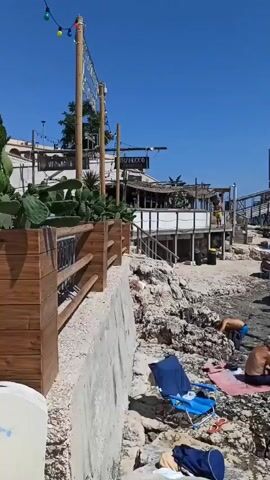 The video shows a rocky coastline on a sunny day. Several people are relaxing on the rocks, some sitting in chairs and others lying on towels. A few people are in the water, swimming. In the background, there is a wooden structure with multiple levels, possibly a restaurant or bar, and some smaller buildings. There are also some plants, including cacti, in wooden planters along a railing. String lights are visible above the railing. A few boats are in the distance on the blue sea. The scene appears to be a beach resort or a popular spot for sunbathing and swimming.