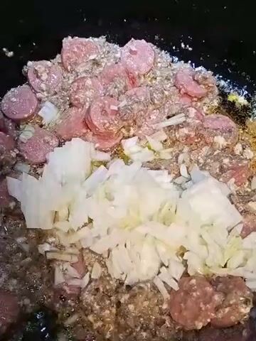 A close-up shot shows a dark cooking pot filled with various ingredients. In the foreground, a large pile of chopped white onions sits on top of other food items. The ingredients underneath the onions include sliced pink sausage and crumbled ground meat. Some pieces of the meat appear to be seasoned with spices. The food is being cooked, evidenced by the shimmering oil and the slightly browned appearance of some of the ingredients. The lighting is somewhat dim, focusing attention on the food within the pot.