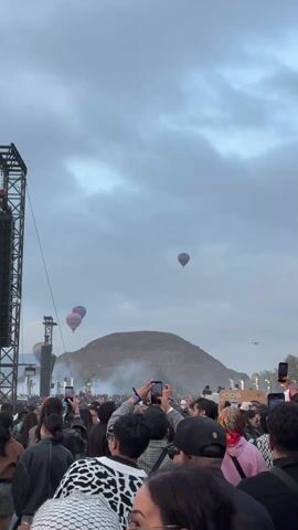 The video shows a large crowd of people facing away from the camera, their attention directed toward hot air balloons floating above what appears to be a large pyramid or hill. The crowd is densely packed, and many individuals are holding up their phones, presumably to capture photos or videos of the spectacle. To the left, a tall, black speaker tower is visible, suggesting the presence of a stage or performance area. The sky is overcast with grey clouds, and some haze obscures the base of the pyramid. A few promotional signs can be seen on the right.