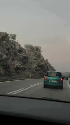 The video shows a view from inside a car driving on a highway. The road is paved and has white lane markings. To the left of the road is a steep, rocky hillside with some sparse vegetation. Several cars are visible on the road, including a turquoise car in the foreground. The sky is overcast and the lighting suggests it is late afternoon or early evening. The car's dashboard is visible at the bottom of the frame.