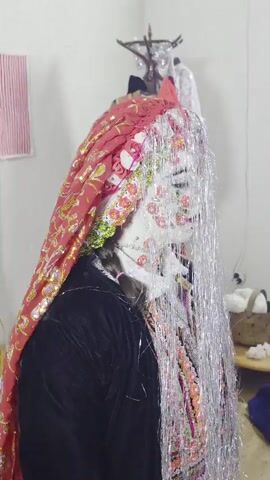 A person in traditional wedding attire, possibly a bride, is shown from the side. They are wearing a white face covering with red floral designs, a red and gold veil, and a dark garment. Silver tinsel hangs down from the head, obscuring the face. The background appears to be a room with clothing on a rack and some decorative items. The person is stationary.