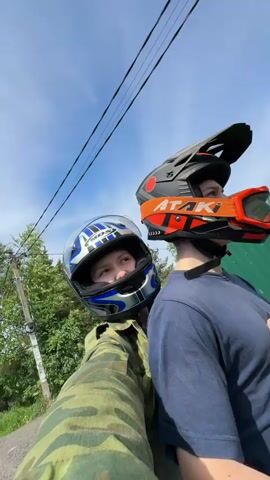 A selfie-style video captures two individuals outdoors. On the left, a woman wearing a camouflage jacket and a silver and blue full-face MDS helmet looks at the camera with a slight smile. She leans against the back of a man on the right, who wears a dark blue t-shirt and an orange and black full-face ATAK motocross helmet with attached goggles. The man faces right, away from the camera. The background features a blue sky with clouds, power lines, green trees, a green fence, and a red building. The sound of an engine idling is present throughout the video.