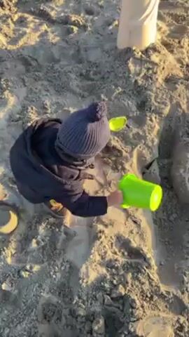 A small child wearing a navy-blue puffy jacket, yellow pants, and brown shoes plays in the sand on a beach.  They wear a navy-blue knit hat with a pompom. The child is filling a bright green bucket with sand, viewed from above.  A portion of a person's leg covered in white fabric is visible to the right of the child. The sand is grey. The light source suggests it's either early morning or late afternoon.