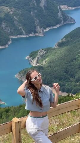 This media file features a young woman with long, brown hair and white sunglasses posing for a photo on a sunny day. She is wearing a light blue, cropped collared shirt and white capri pants with a green belt. The woman is standing behind a wooden fence, leaning on it with her left arm. Her right hand is near her hair, and she is smiling at the camera, looking happy and relaxed.

The background is a breathtaking natural landscape. A vast, winding river with bright blue water flows through a deep canyon. The canyon walls are steep and covered in lush green trees and rugged, rocky terrain. The overall scene suggests a scenic overlook, possibly in a mountainous region. The lighting indicates it is daytime, likely mid-morning or afternoon, with clear, bright conditions. The abundance of green foliage suggests a warm season, possibly summer or late spring. The location is identified as Dubljevići, Montenegro, known for its dramatic natural beauty. There is no visible text in the media file.
