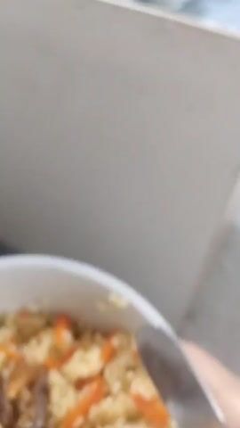 The video shows a bowl of pilaf held by a person's hand. The pilaf consists of rice, sliced carrots, and a piece of meat. A metal spoon is also visible near the edge of the bowl. The background shows a white or light-colored wall and a glimpse of what appears to be a staircase. The recording seems to have been captured indoors. Sounds of people chattering can also be heard in the background.