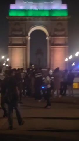 The video shows the India Gate monument illuminated at night with the colors of the Indian flag: orange at the top, white in the middle, and green at the bottom. The monument is lit with purple lights along its lower arches. A crowd of people are gathered in front of the monument, some appearing to be tourists or locals enjoying the evening. A few individuals are walking, while others are standing and observing the monument or interacting with each other. One person in the foreground is holding a small blue drone. The environment appears to be a public space with streetlights visible in the background and vehicles in the distance. The overall atmosphere seems to be festive and celebratory.