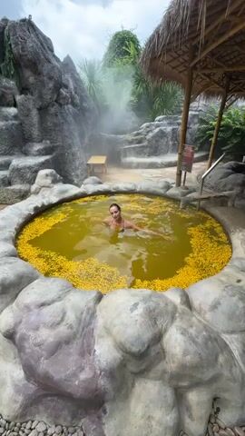 A woman is in a circular stone hot tub filled with yellow flowers. She is wearing a black bikini. Steam rises from behind the tub. The hot tub is surrounded by rock formations and tropical vegetation. There is a small wooden structure with a thatched roof next to the tub. The sky is overcast. The hot tub is made of grey stones with a rough, uneven surface. The water is yellowish-brown in color. The woman turns her back to the camera then floats for a moment.