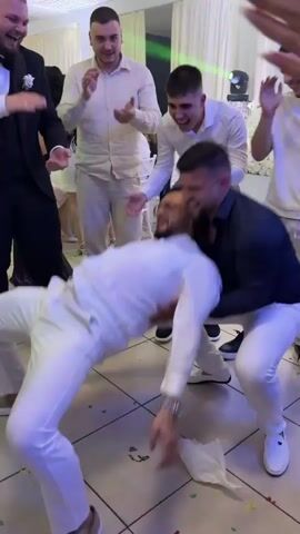 The video shows a group of men at what appears to be a wedding reception, based on attire and celebratory atmosphere. One man in a white suit is being playfully held up by another man, while others clap and laugh around them. The group is in a banquet hall with tiled floors and white drapery. There's confetti on the floor. Lighting includes overhead lights and stage lighting in green and blue. Some men are wearing formal attire such as suits or dress shirts.