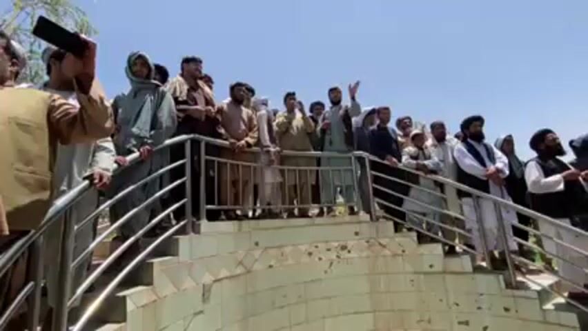 Here's a detailed description of the video:

**Scene Overview:**
The video shows a group of men standing on a tiered platform or structure, likely overlooking something that isn't visible in the frame. The scene is outdoors, with a clear blue sky visible.

**People:**
*   The group consists entirely of men.
*   They are wearing a variety of clothing, including traditional garments such as shalwar kameez and turbans. Some are wearing vests or jackets over their clothing.
*   Some men have beards.
*   One man on the left side is holding a cell phone and appears to be recording the scene.
*   Most of the men are looking in the same direction.

**Objects:**
*   A set of stairs and metal railings.
*   A structure underneath the stairs with pale green tiles.
*   Cell phone being held by one of the men.

**Actions:**
*   The group of men are observing something that is occurring off-camera.
*   At least one man is recording with his phone.
*   Some men appear to be talking or gesturing, but overall the atmosphere seems relatively calm and observant.

**Environment:**
*   The weather appears to be sunny and clear.
*   There is some greenery visible in the background, suggesting the location is outdoors in a possibly rural or semi-rural area.

**Overall Impression:** The video captures a moment where a group of men are observing or documenting something.