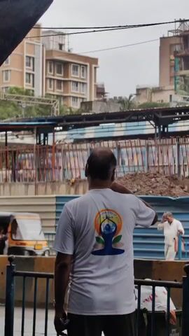 A bald man in a white t-shirt with a colorful graphic on the back stands facing away from the camera, looking towards a construction site. In the background, there are several buildings under construction, some with scaffolding. A yellow auto-rickshaw and other vehicles are visible on the road in front of the construction site. The sky is overcast.