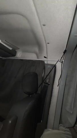 The video shows the interior of a vehicle, possibly a van or camper. The camera pans from the ceiling down to the seats and dashboard. There are dark-colored curtains hanging from the ceiling, dividing the interior. One of the curtains is slightly open, revealing some belongings underneath. A seatbelt is visible, as well as part of a seat. The dashboard has buttons and a red light on. The overall impression is of a functional, customized interior space.