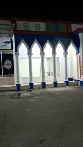 A person is sleeping on the ground near the entrance of a building. The building has an arched entryway and is lit by overhead lights. The surrounding area is paved and dark, suggesting it is nighttime.