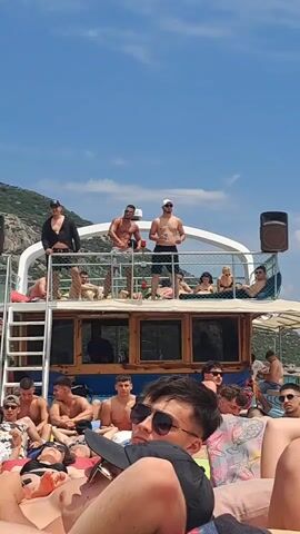 This video captures a lively daytime boat excursion in Menderes, Turkey, against a clear blue sky and a backdrop of green, hilly coastline. The scene appears to be a boat party or a relaxed trip on a two-story vessel, filled with people.

On the upper deck, several young men are visible, some shirtless, while others wear caps and light clothing. One man on the left, wearing a black shirt and cap, is seen raising his arm. Another shirtless man in the center looks towards the right, and a third shirtless man to his right holds a red cup. A large speaker is positioned on the right side of the upper deck, and a "GARMIN" device is mounted in the center. Other individuals are seen relaxing and lounging on this deck.

The lower deck is also crowded with people, mostly young men, many of whom are shirtless or wearing t-shirts and sunglasses. They are shown relaxing, sitting, and lying down. In the foreground, a man wearing a black cap and sunglasses is prominently featured, taking a drink from a red cup. The overall ambiance is casual and celebratory, indicative of a fun day out on the water under sunny weather conditions.