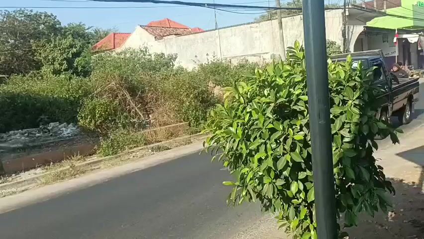 The video shows a black minivan driving down a paved road, partially obscured by a large green bush in the foreground. The van has "FAHA" written on its side. Further down the road, two people ride a black motorcycle. The person in front is wearing a black hoodie and shorts, while the person behind is wearing a grey hoodie and shorts. The environment appears to be a rural or suburban area with houses, trees, and a white wall visible in the background. The sun is shining, indicating it is daytime.