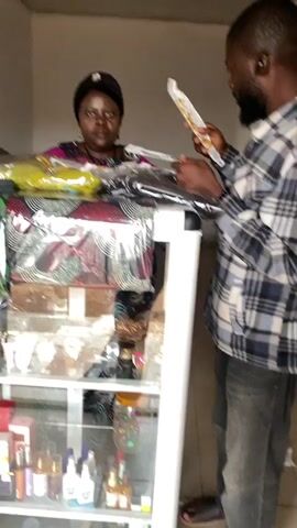 The video shows a retail display case with two individuals standing near it. A woman, wearing a headscarf and a colorful top, is positioned behind the display case. She is looking towards the right side of the frame and appears to be speaking or gesturing with her hands. A man, wearing a plaid shirt, stands to the right of the display case, facing the woman. He is holding a long, thin object, possibly a product or receipt, and looking at it. The display case is made of glass and metal and is filled with various items, including watches on the upper shelf and perfume bottles and boxes on the lower shelves. The background appears to be an indoor setting with plain walls. The lighting is somewhat dim, with some shadows present. The video is shaky and appears to be handheld.