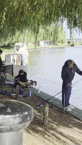 A group of people are fishing by a river. The person in the foreground is fishing with two rods. Behind them, two other people are sitting and fishing. In the background, there are boats moored along the riverbank. The area is covered in trees, providing shade. The weather appears to be sunny.