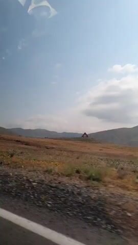 A vertical, shaky video shows a landscape with hills, fields, and a clear blue sky with some clouds. The foreground has a dirt road and some dry grass. In the distance, there are more hills and a small house with a red roof. The sun appears to be shining. The video pans slightly to the right, giving a sense of movement, possibly from a vehicle. The overall impression is of a rural, natural environment.