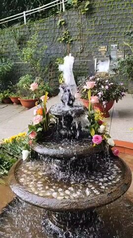 A three-tiered fountain with water cascading down each level is the central focus of the video. The fountain is decorated with a variety of pink and white roses, as well as some orange and yellow flowers. The fountain is surrounded by small, smooth pebbles. In the background, there is a stone wall covered in ivy, as well as potted plants and flowers. The lighting appears to be natural, suggesting an outdoor setting. There are no people visible in the video. The overall scene evokes a sense of tranquility and natural beauty.