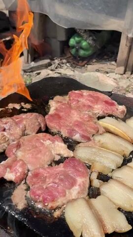 The video shows pieces of raw meat and fatty strips of meat being cooked on a flat, dark cooking surface. The meat is pink and raw in some areas and appears to be sizzling on the hot surface. In the background, there are flames from a fire, some brick-like structures, and a green object that appears to be a bell pepper. The overall environment suggests an outdoor cooking setup.