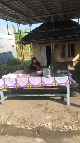 A woman wearing a black headscarf and a maroon shirt sits behind a table laden with food. She is holding a utensil, possibly a spoon, and appears to be interacting with the food or a bowl in front of her. The table is covered with a purple and yellow patterned cloth and displays various items including bowls, a pot, and what appear to be food containers. In the background, a simple house with a dark roof is visible, partially obscured by a covered seating area. The environment seems to be outdoors, with rough ground and some vegetation suggesting a rural or semi-urban setting. The lighting suggests it is daytime.