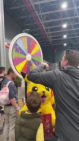 The video shows a crowd of people standing around a brightly colored wheel and a large yellow Pikachu statue at what appears to be an event or convention. The wheel is spinning rapidly, displaying various colors and possibly letters or symbols. A young boy in a vest is positioned directly in front of the Pikachu, looking at it intently. Several adults are nearby, observing the scene. One adult wears a blue shirt and a pink backpack, and another is in a grey jacket. The environment is a large, open space with a high ceiling and visible structural elements like pipes and beams. There is a sign beneath the Pikachu, possibly related to a game or prize. The overall atmosphere seems casual and engaging, with people interested in the spinning wheel and the Pikachu.