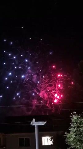 This is a vertical video of fireworks exploding in the night sky. The fireworks are primarily red and blue, with some pink and purple hues. There are also some visible trails of smoke from the explosions. In the foreground, there is a dark silhouette of trees on the right side of the frame, and some power lines and a building or structure are visible at the bottom. A street light fixture is also present in the lower center of the frame. The sky is completely dark, indicating it is nighttime.