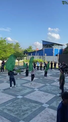A group of young people are practicing a flag performance outdoors on a paved area with a checkered pattern. Several individuals in the foreground are holding large green flags, some of which are in motion. In the background, another group of people appear to be holding musical instruments, suggesting a marching band practice. The sky is blue with some scattered white clouds. Behind the participants, there is a blue building with a large white structure on its roof, and some trees and foliage. The lighting suggests it is daytime, likely morning or afternoon.