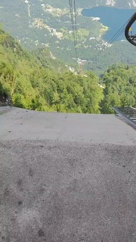 The video is a POV shot from a cable car ascending a mountain. In the foreground, there is a concrete path leading to the edge of the cable car station. Beyond that, the cable car wires stretch upwards towards the sky. In the distance, there is a panoramic view of a valley with a lake, lush green trees, and a small village nestled amongst the mountains. The sky is partly cloudy, and the overall impression is one of breathtaking natural beauty.