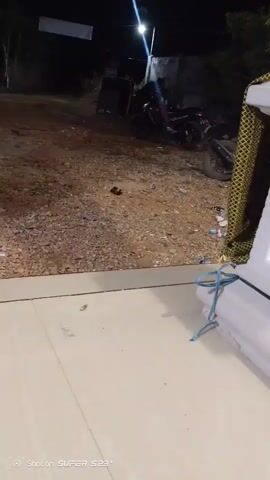 The video shows an outdoor scene at night. In the foreground, there is a white object with a yellow and black patterned covering on the right side, and it is tied with a blue rope. The ground in the foreground is paved with light-colored tiles. In the background, there is a dirt or gravel area with scattered debris. A street light illuminates the area. A red motorcycle is parked behind a large black speaker. There appears to be a person near the motorcycle, but they are not clearly visible. A white banner is hanging in the upper left corner of the frame. The overall impression is that of an outdoor event or gathering area at night, possibly in a rural or less developed setting.