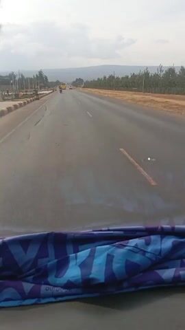 The video is taken from inside a moving vehicle on a highway. The road is asphalt with some fading orange lane markings. There is sparse traffic on the road including a motorcycle and van in front of the camera. Beyond the road are small trees. In the distance is a long, low mountain range under a cloudy sky. The dashboard is visible at the bottom of the frame, covered with a blue patterned cloth.
