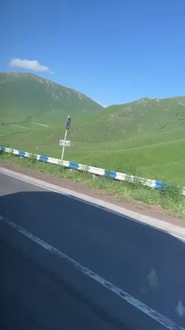 This media file captures a scenic drive through a lush, mountainous landscape. The scene is dominated by rolling green hills under a clear blue sky, suggesting a pleasant, sunny day. A paved road with white lane markings curves through the terrain, bordered by a guardrail with white and blue accents.

Visible on the side of the road are two traffic signs. The first sign, partially obscured by the guardrail, appears to be a speed limit sign indicating "40". Below it, another sign displays "121". The second sign, appearing further down the road, is a warning sign with a red border and a symbol that depicts a sharp turn or series of turns.

The camera appears to be mounted on a moving vehicle, giving the viewer a sense of traveling through this picturesque environment. There are no people visible in the frame. The overall impression is one of a peaceful journey through a natural, undeveloped area, likely in a rural or mountainous region. The time of day appears to be daytime, given the bright sunlight and blue sky. The location context provided, Tsilkar, Armenia, further supports the interpretation of a mountainous, possibly rural, setting.