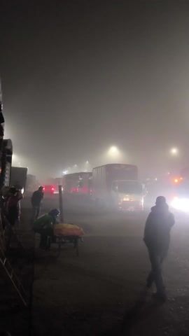The video shows a very foggy scene, likely at night. Several trucks and cars are present, illuminated by their headlights and streetlights, which create a bright glare through the fog. Several people are visible, some standing near the vehicles and others walking. One person in the foreground is walking toward the vehicles, and another person can be seen in the bottom right corner of the frame. The ground appears to be pavement or a similar hard surface.