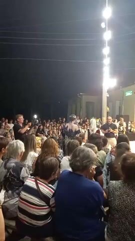A crowd of people watches a performance at night. String lights are visible overhead. In the center of the frame, a performer in a black and white dress and a head covering dances while another person in a white head covering and patterned outfit moves to the side. A man in a black shirt films the performance with a camera. Another man in a blue shirt and white shorts stands to the right, gesturing. The audience is seated and standing, facing the performers. The background shows buildings and trees.