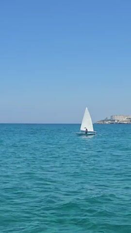 A sailboat with a white sail is gliding across the bright blue water. A person is standing in the sailboat. The sky is clear and blue. Buildings are visible on the horizon.