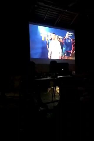 The video shows a projection screen in a dark room. On the screen, there is an image of a man in a white suit, and behind him, several other people are visible. In the foreground, there are silhouetted figures of people sitting at tables. The room appears to be set up for watching a presentation or movie. The projector is not visible in the frame. The quality of the video is low, and the lighting is dim.
