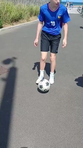 A young person wearing a blue soccer jersey with the number 10 and black shorts is practicing with a soccer ball on an asphalt surface. The person kicks the ball multiple times, appearing to be performing footwork drills. In the background, there is vegetation, a sidewalk, and other people walking. The day appears to be sunny, casting shadows on the ground.