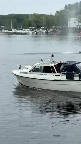 A white motorboat with a blue stern canopy travels from right to left across a calm body of water. The boat features a small cabin, antennae, a radar dome, and a visible registration number "M-12479." Fishing nets and a red life buoy are mounted on the stern. In the background, a marina with numerous docked sailboats and motorboats lines a distant shore covered with green trees under an overcast sky. The boat generates small ripples in the water as it moves.