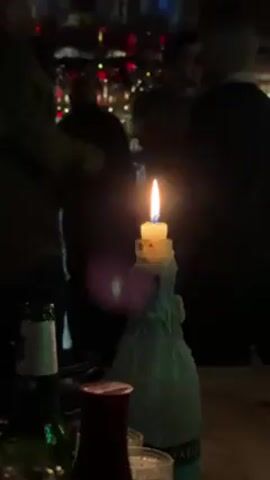 A dimly lit, tight scene shows a candle burning on top of a bottle with wax dripped down its sides, on a wooden table surrounded by people with bottles and other drinking glasses. The background is blurry but shows lights, some red, strung up. Most of the people are silhouetted as if facing away from the camera; a couple have unclear faces. The atmosphere seems lively.