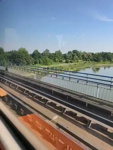 A train moves along tracks next to a body of water and a bridge. The sky is clear and blue with some clouds. The surrounding area is green with trees and vegetation. The video is filmed from inside the train, looking out the window.