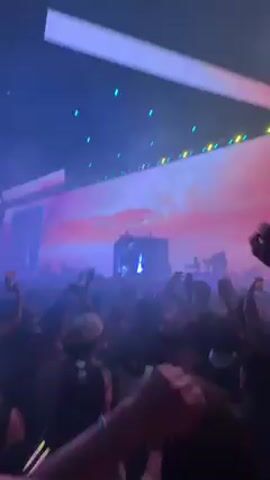 A dynamic low-angle shot captures a lively indoor music concert in Palm Desert, United States. A large crowd of people is seen from behind, with many raising their hands and recording the performance on their phones, indicating an enthusiastic atmosphere.

In the center of the stage, a DJ booth is visible, where a DJ is performing. Behind the DJ, large screens display vibrant, abstract visuals dominated by hues of pink, purple, and blue, resembling clouds or a surreal sky. Bright spotlights and stage lighting in blue and white illuminate the venue, casting a hazy glow through what appears to be smoke or fog effects. Long, rectangular light fixtures are positioned above the stage. The overall ambiance is energetic and immersive, characteristic of a live music event.