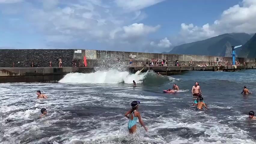 The video captures a bustling scene at a rocky beach with a concrete pier. The sky is partly cloudy, and a mountainous landscape is visible in the background. Numerous people are enjoying the water and the pier. In the foreground, a young person in a blue swimsuit and goggles stands in shallow water, facing away from the camera. To their left, a young person with dark hair and goggles runs out of the water. Several other children are playing in the surf, some with bodyboards. A large wave is breaking, generating white foam. On the pier, many people are gathered. Some are sitting, others are standing, and a few are interacting with the water. Red and orange surfboards lean against the pier's wall. The pier itself is lined with graffiti. The overall atmosphere is lively and recreational.