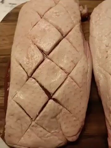 The video shows three pieces of raw duck breast, each with its skin scored in a diamond pattern, resting on a dark wood cutting board. The surface the board is sitting on is white with a muted geometric pattern. The pieces of duck breast are pinkish-white in color, and small pools of liquid have collected in the scored lines of the skin. The camera pans slightly to the left, then zooms out slightly, revealing the third piece of duck breast on the left side of the frame. The lighting is soft and diffused.