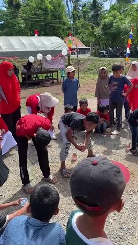 The video shows an outdoor event with several children and adults. Many of the children are participating in a game, bending over to interact with objects on the ground. One child in the foreground is wearing a red baseball cap and looking to the right. To the left, a woman in a red hijab and red shirt is also looking down. Other children in red and white outfits are also bending over. In the background, there are tents, trees, and several motorcycles. Colorful flags are visible in the background. The ground is covered with gravel. The event appears to be festive, possibly a community gathering or competition.